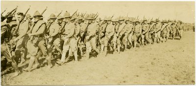 Horizontal, sepia photograph showing rows of uniformed soldiers marching through a grass field in formation while carrying rifles by Carl Michel